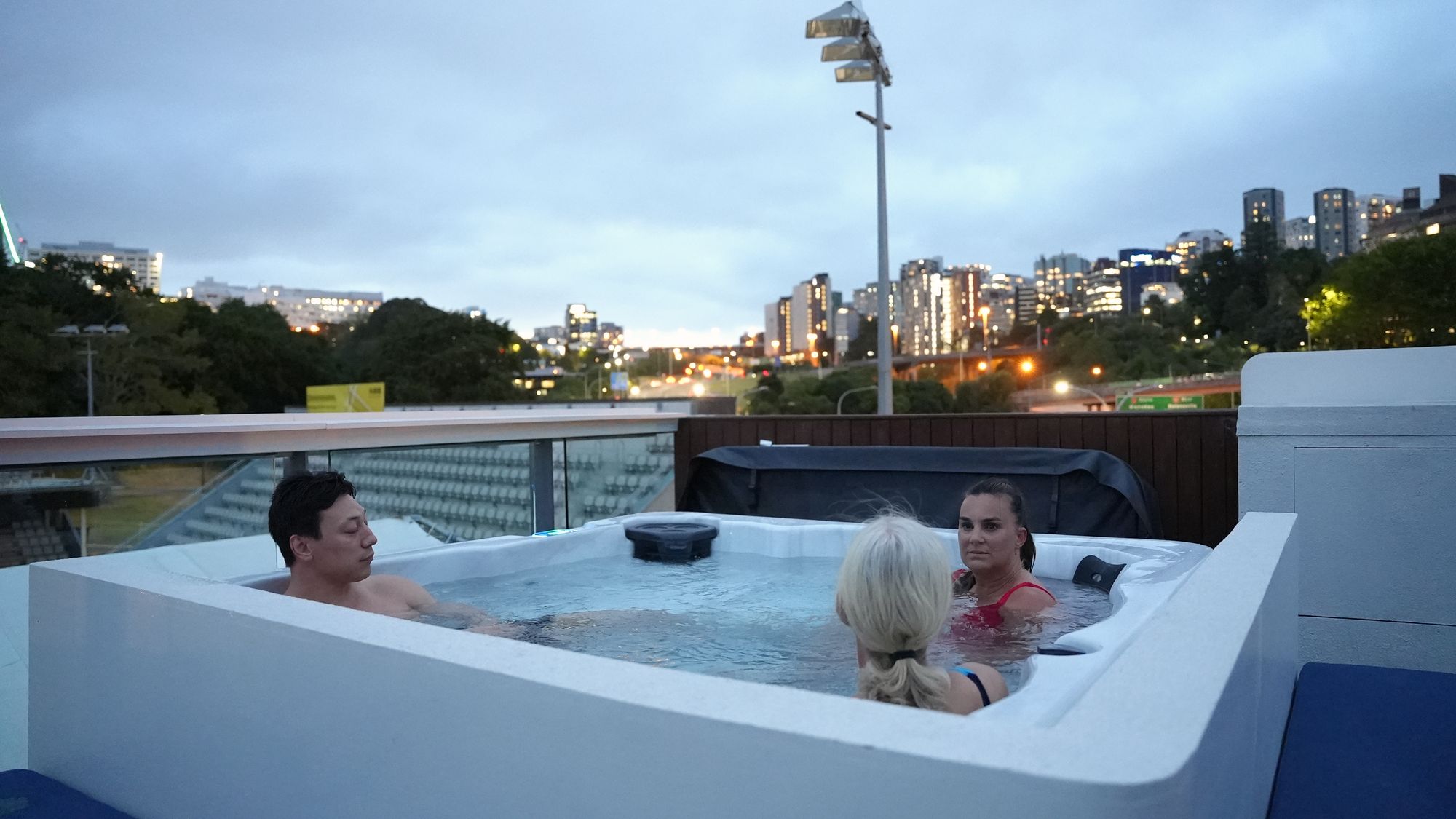 Three people in the rooftop spa bath at Renew at dusk, with the Auckland skyline behind them