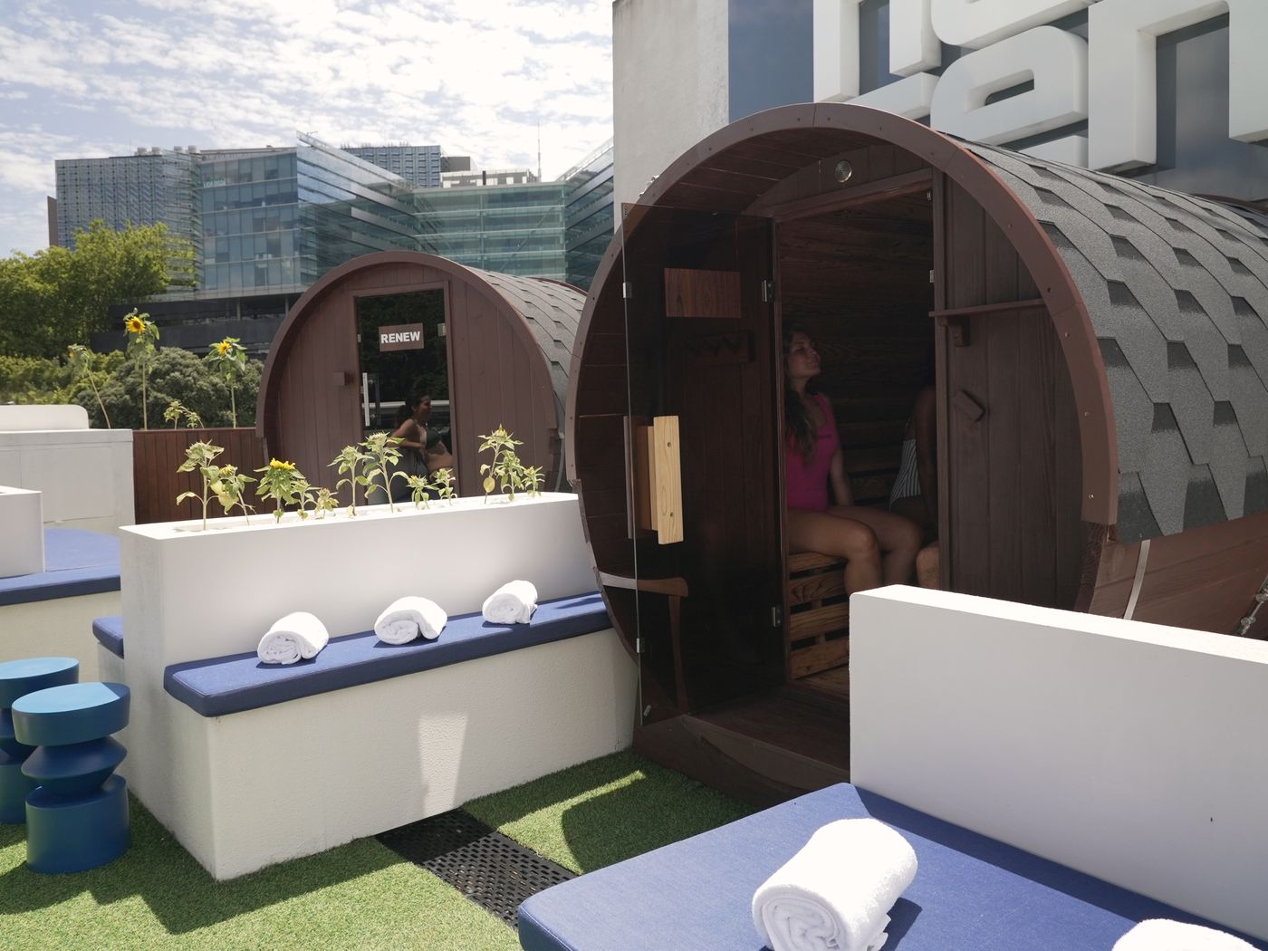 The two barrel saunas on the Renew rooftop, one with the door open and someone sitting inside, with rolled towels on the bench and the Auckland skyline behind